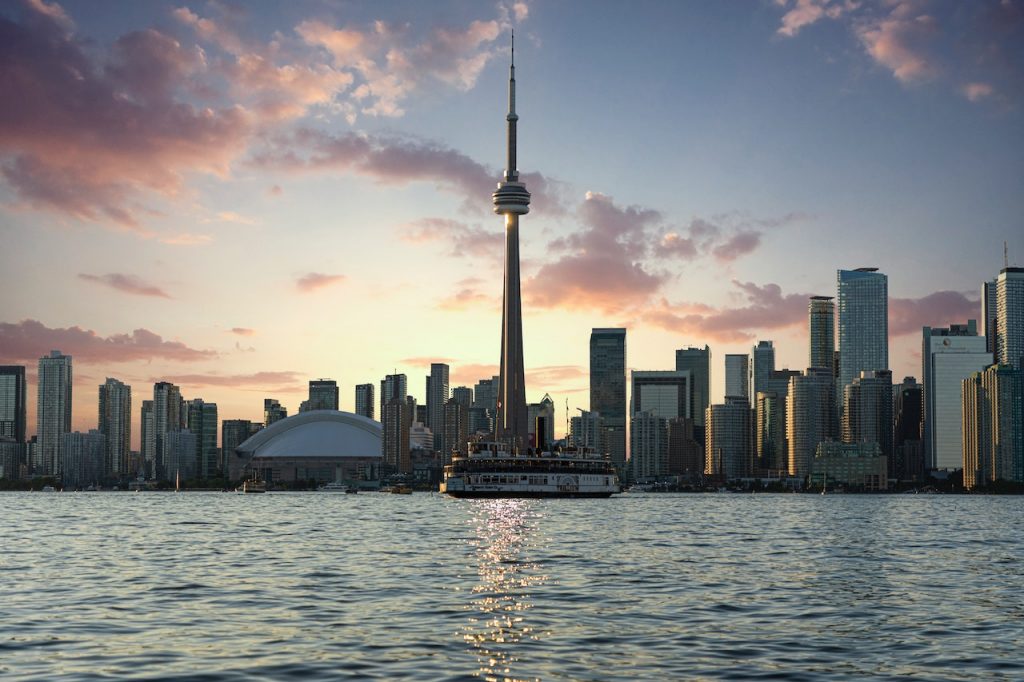 A photo of the Toronto skyline at sunset, with the CN Tower prominently featured. The sky is lit up in shades of orange and pink, creating a beautiful backdrop for the cityscape.